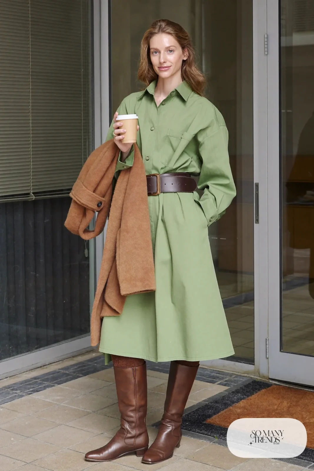Woman in pine green belted shirtdress, black tights, chocolate boots and wool coat over her arm, outfit 17