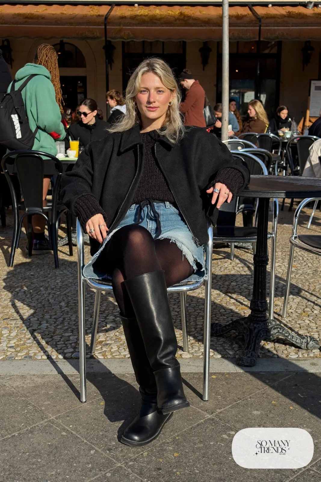 Woman in a cropped black jacket, chunky knit, faded denim cut-off shorts over black tights and tall black boots, outfit 12