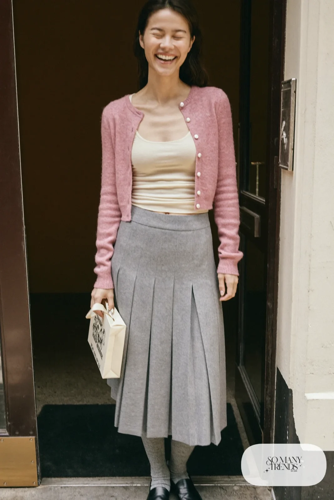 Woman in a dusty pink cropped cardigan, cream tank, gray pleated midi skirt, gray tights and black loafers, outfit 7