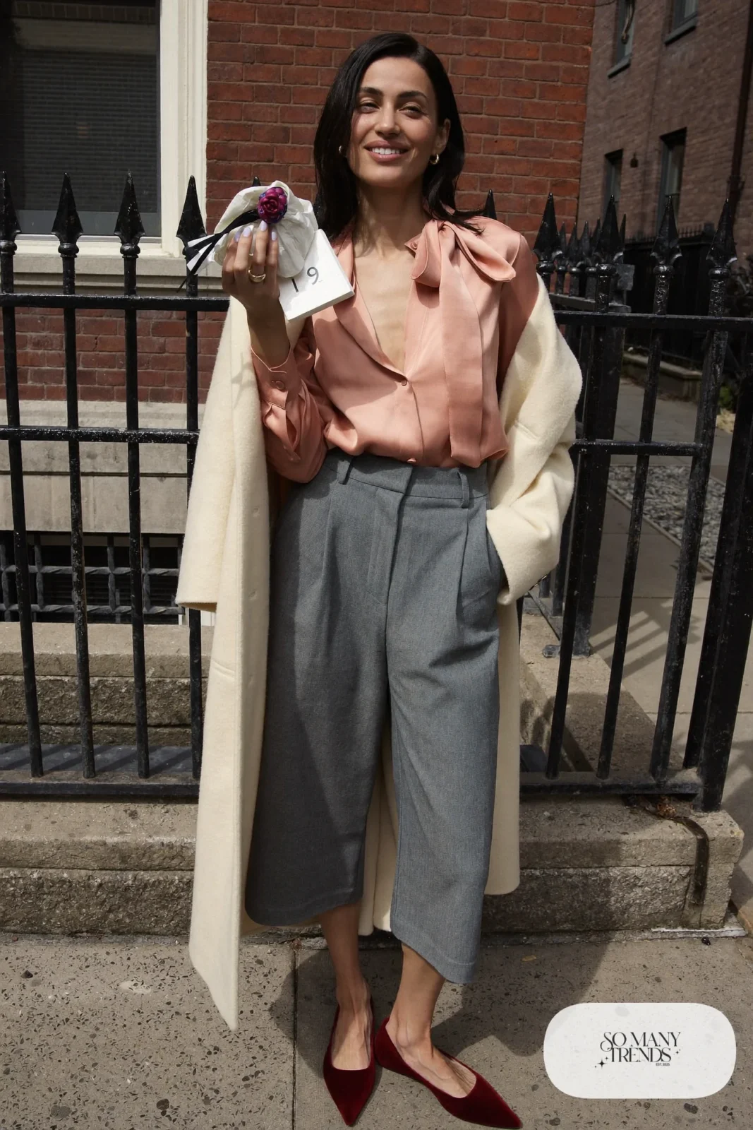 Woman in a pink bow-neck blouse, gray wide-leg crepe pants, burgundy pointed flats and a cream coat, outfit 18