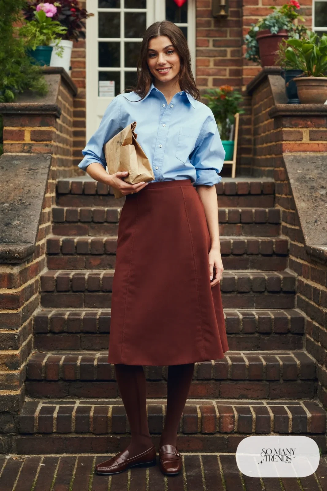 Woman in a soft blue oxford shirt, burgundy A-line midi skirt, black opaque tights and brown loafers, outfit 23