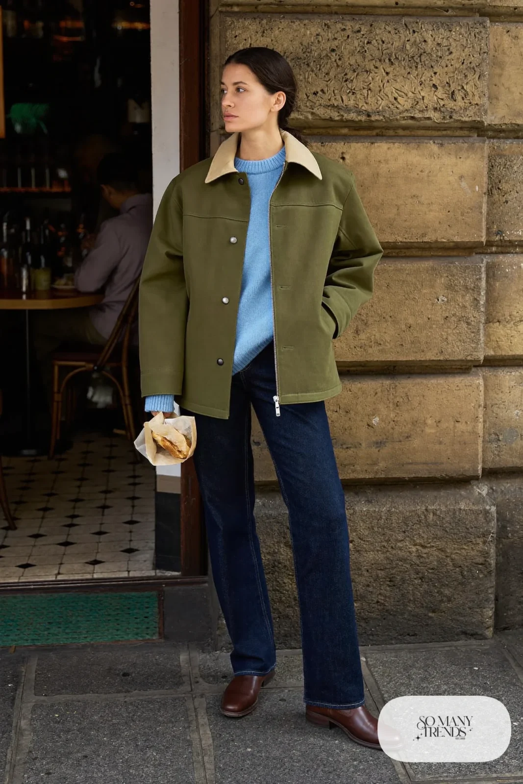 Woman in a baby blue crew-neck sweater, dark straight-leg jeans, olive waxed jacket and brown leather boots on a stone street in Edinburgh, outfit 3