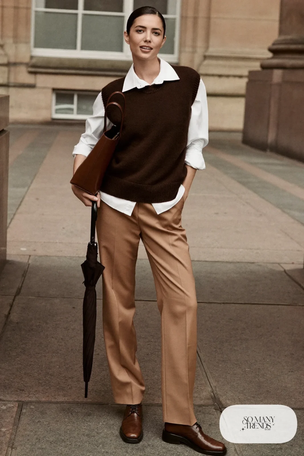 Woman in sand tailored wool pants, white collared shirt, brown knit vest, black wool coat over arm and brown loafer-style boots in a museum courtyard, outfit 4