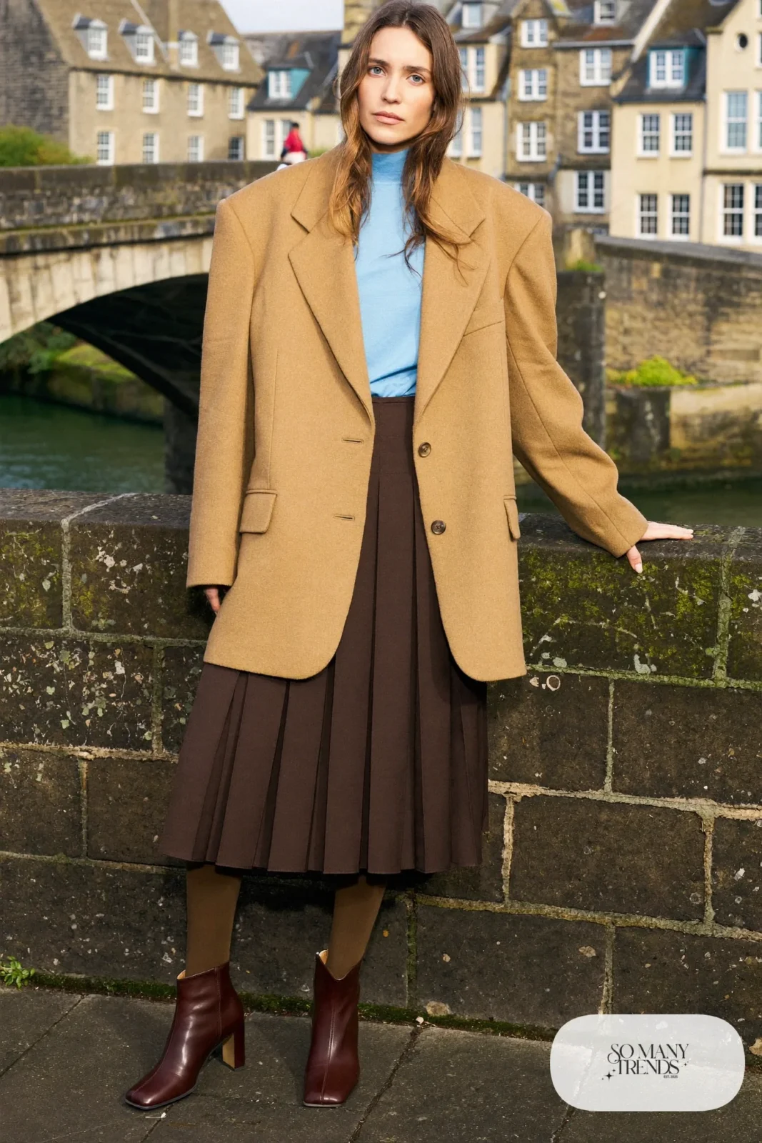 Woman in a camel relaxed blazer, baby blue turtleneck, chocolate brown pleated midi skirt, dark tights and block-heel boots on Edinburgh's Dean Village bridge, outfit 6