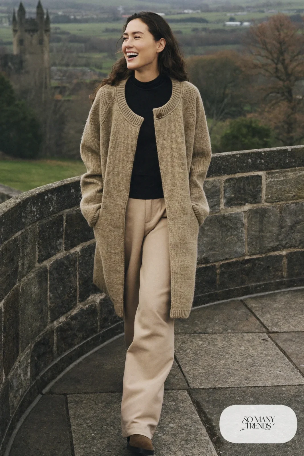 Woman in a long oatmeal knit cardigan coat, black turtleneck, sand wide-leg pants and brown suede boots on castle grounds, outfit 8