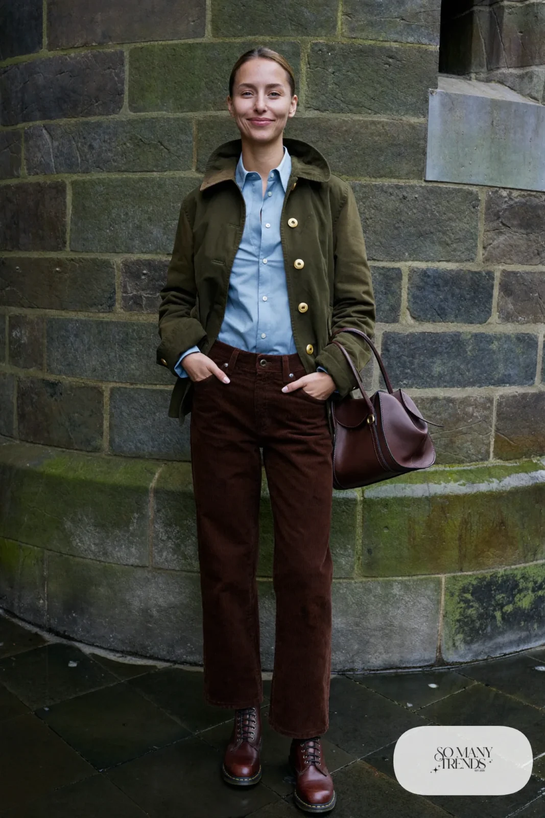 Woman in an olive waxed jacket, baby blue oxford shirt, chocolate corduroy straight pants and brown boots by a castle wall, outfit 10
