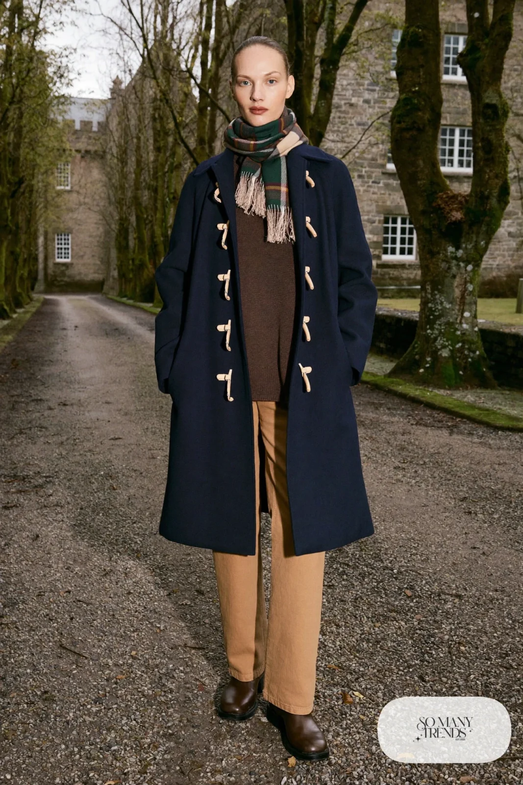 Woman in a navy toggle duffle coat, chocolate brown roll-neck sweater, sand straight pants and brown boots near a castle driveway, outfit 12