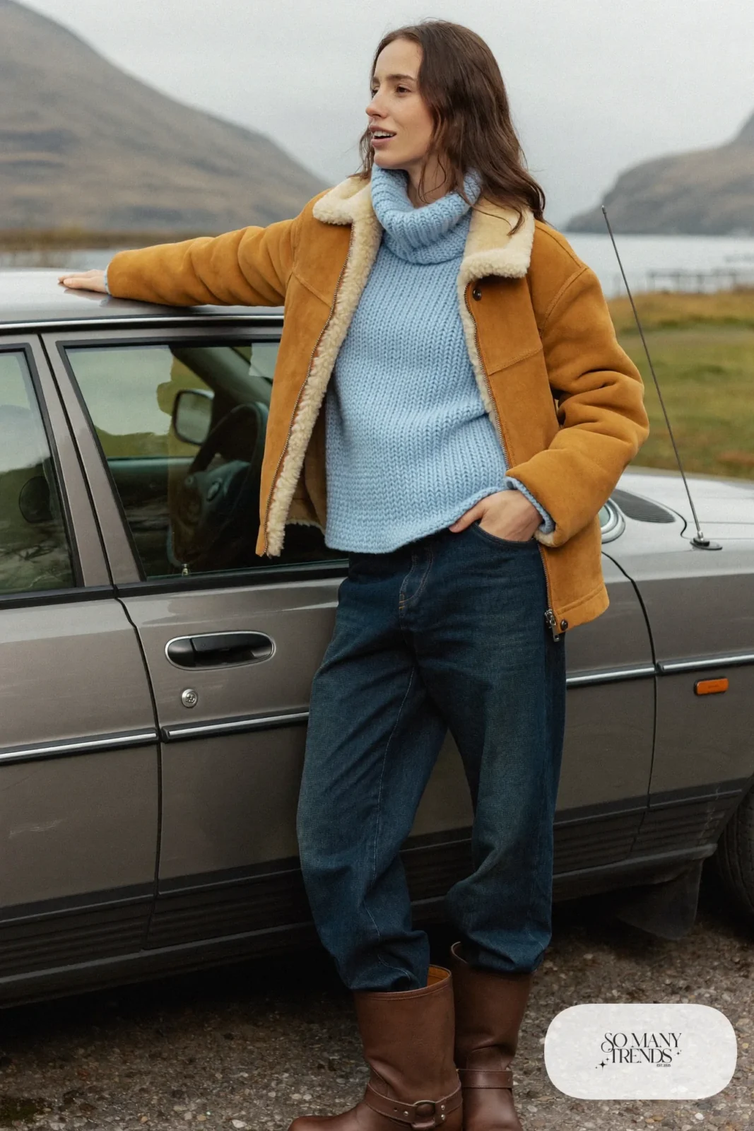 Woman in a sand shearling-lined jacket, baby blue knit sweater, dark jeans and waterproof brown boots beside a car on a Highland road, outfit 13