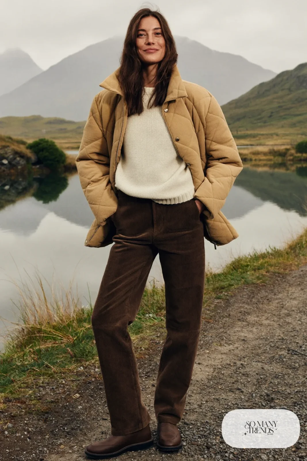 Woman in a sand quilted jacket, cream knit sweater, chocolate corduroy pants and brown waterproof boots beside a loch, outfit 15