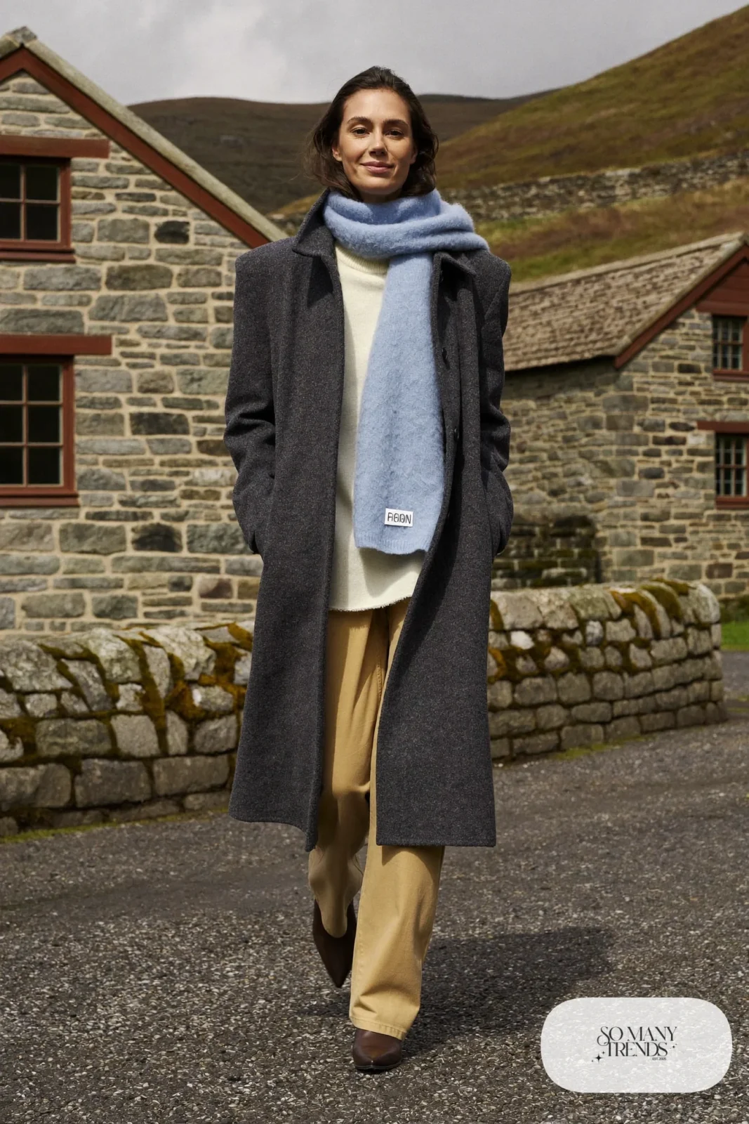 Woman in a charcoal long wool coat, baby blue wool scarf, cream turtleneck, sand pants and brown boots on a Highland village street, outfit 16