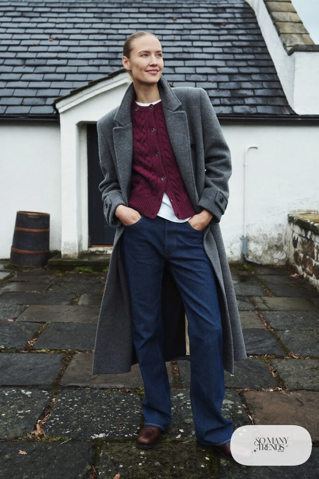 Woman in a heather plum cable cardigan, white tee, dark jeans, long gray coat and brown boots outside a Highland distillery, outfit 17