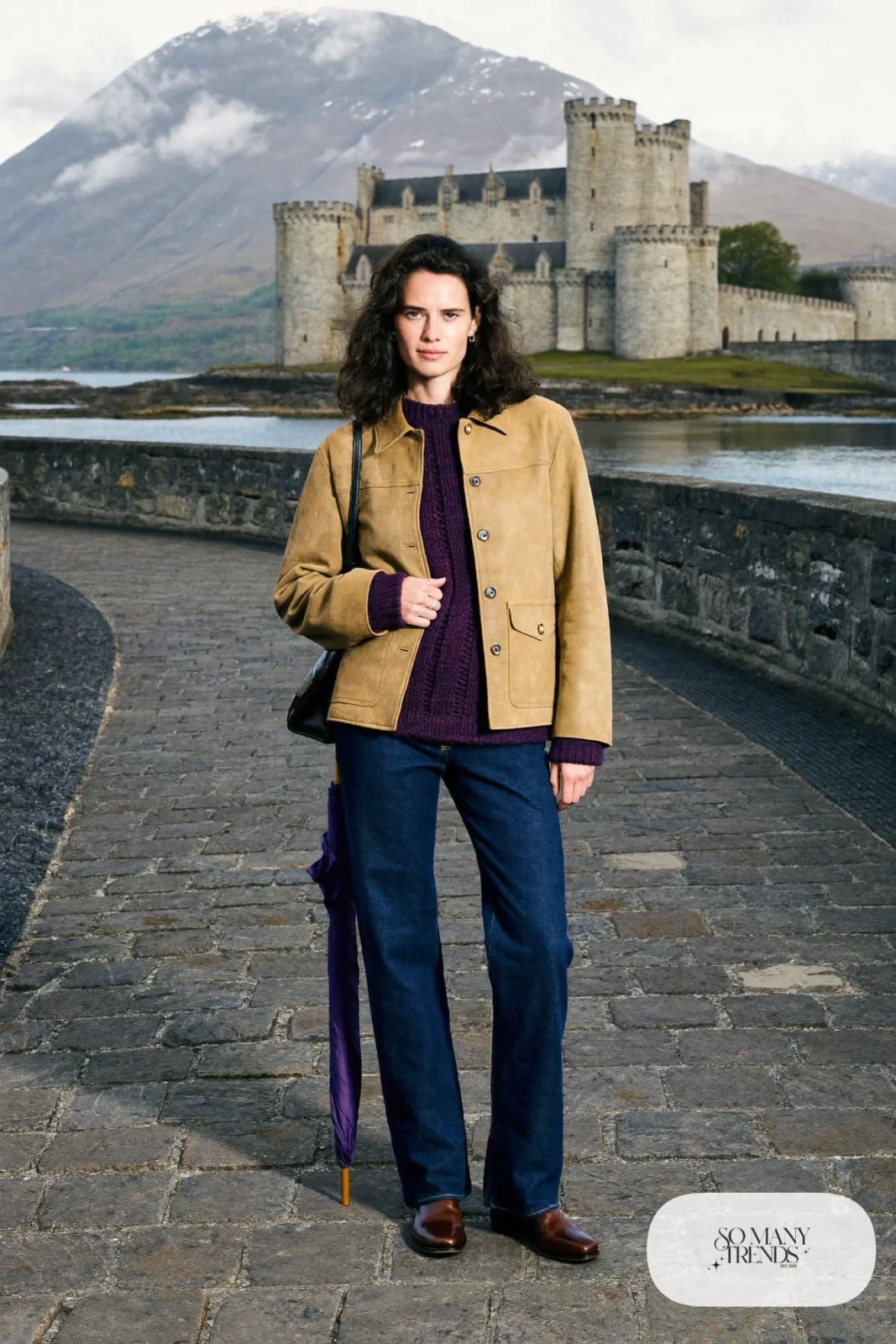 Woman in a sand suede jacket, heather knit sweater, dark jeans and brown boots on a stone causeway at a Highland castle, outfit 19