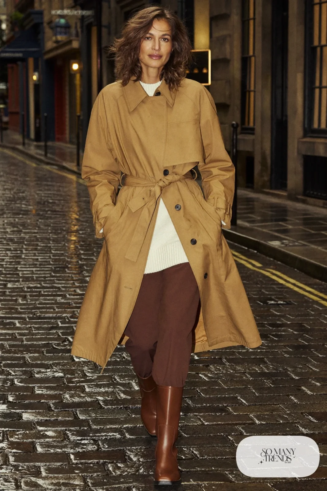 Woman in a sand belted trench coat, cream knit sweater, brown pants and rubber-soled boots on a wet Edinburgh street, outfit 20