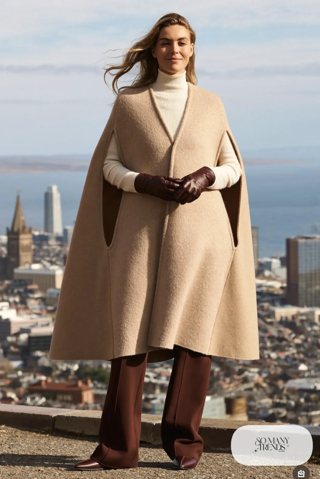 Woman in a sand wool cape with arm slits, cream turtleneck, chocolate wide-leg pants and brown boots on a windy Edinburgh hill, outfit 22