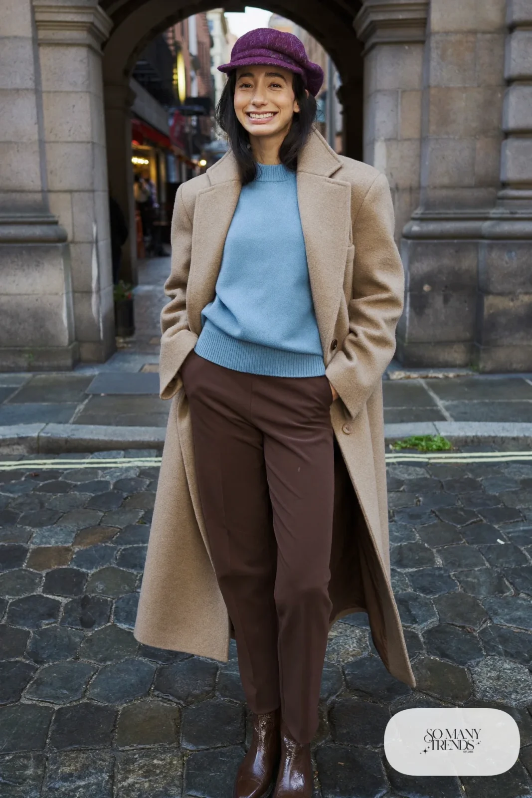 Woman in a plum wool beret, long camel coat, baby blue knit sweater, brown straight pants and brown boots on a rainy street, outfit 23