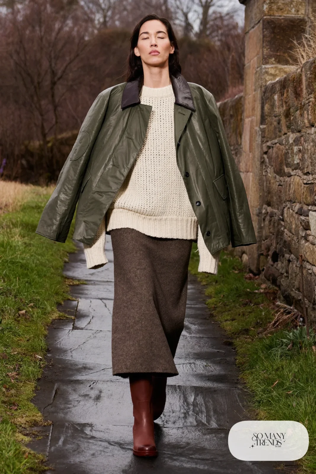 Woman in an olive waxed jacket, cream sweater, gray flannel midi skirt, sheer tights and tall brown boots on a village path, outfit 24