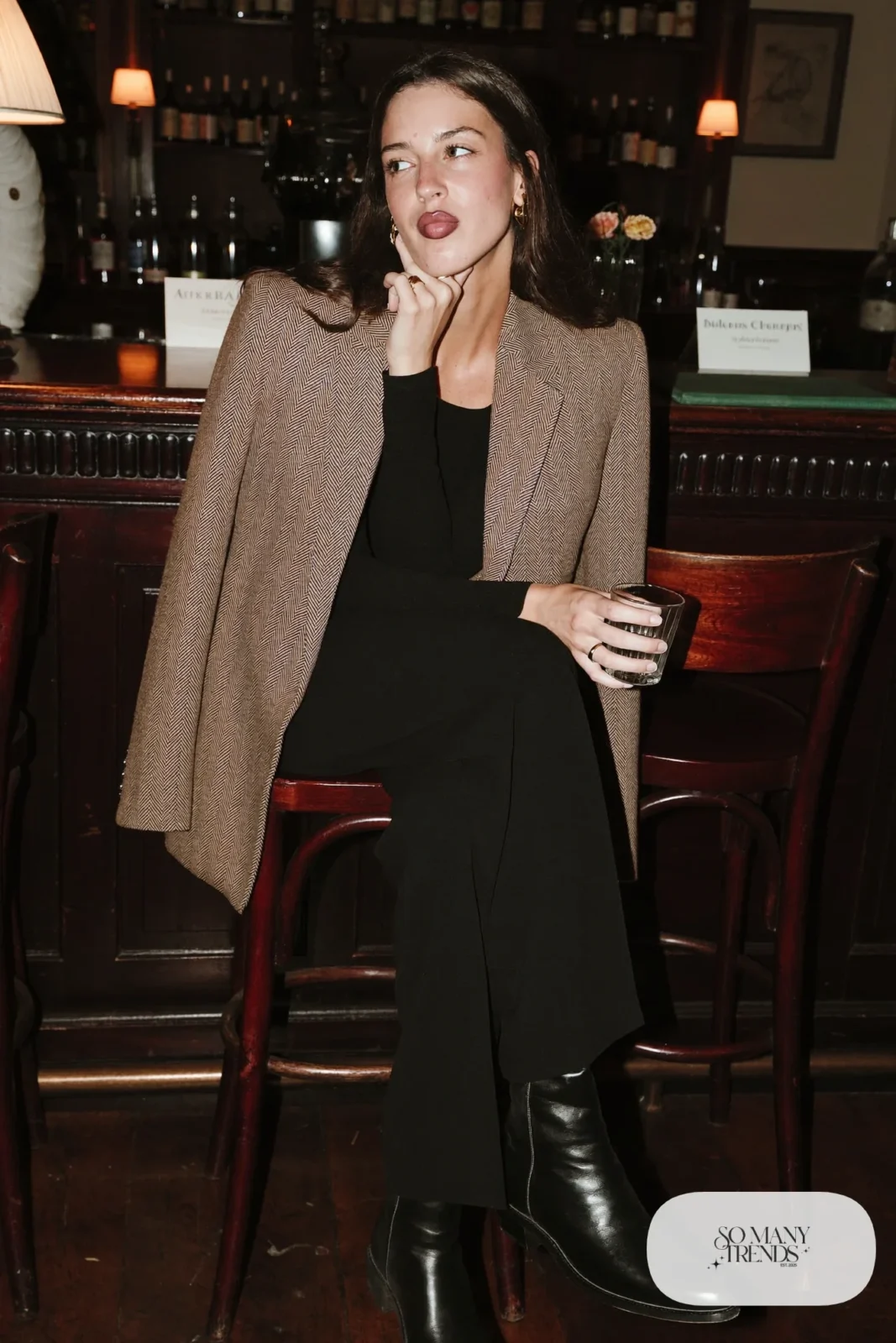 Woman in a brown herringbone tweed blazer, black top, black wide-leg pants and black ankle boots in a whisky bar, outfit 25