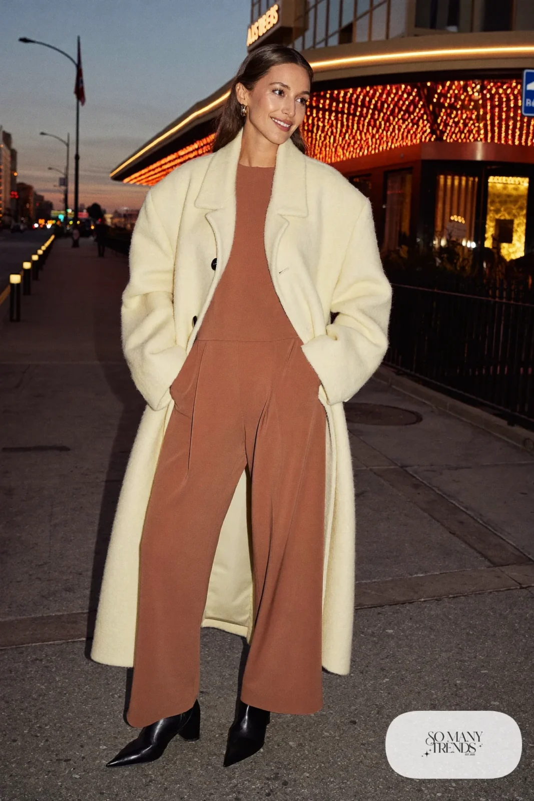 Woman in a terracotta wide-leg jumpsuit, cream long coat and black pointed boots in Las Vegas at night, outfit 10