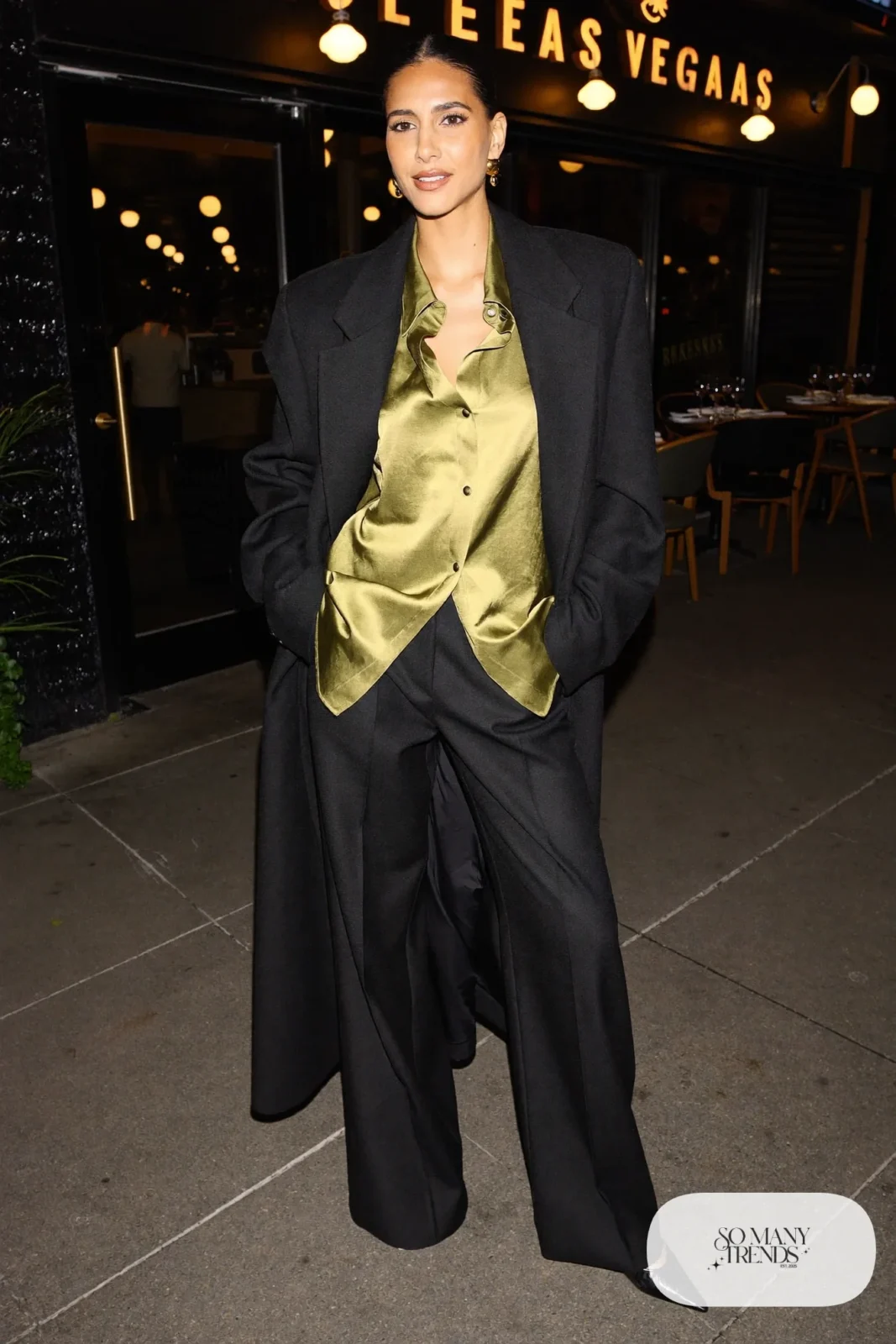 Woman in an olive satin blouse, black wide-leg wool pants, black coat and pumps in Las Vegas at night, outfit 13