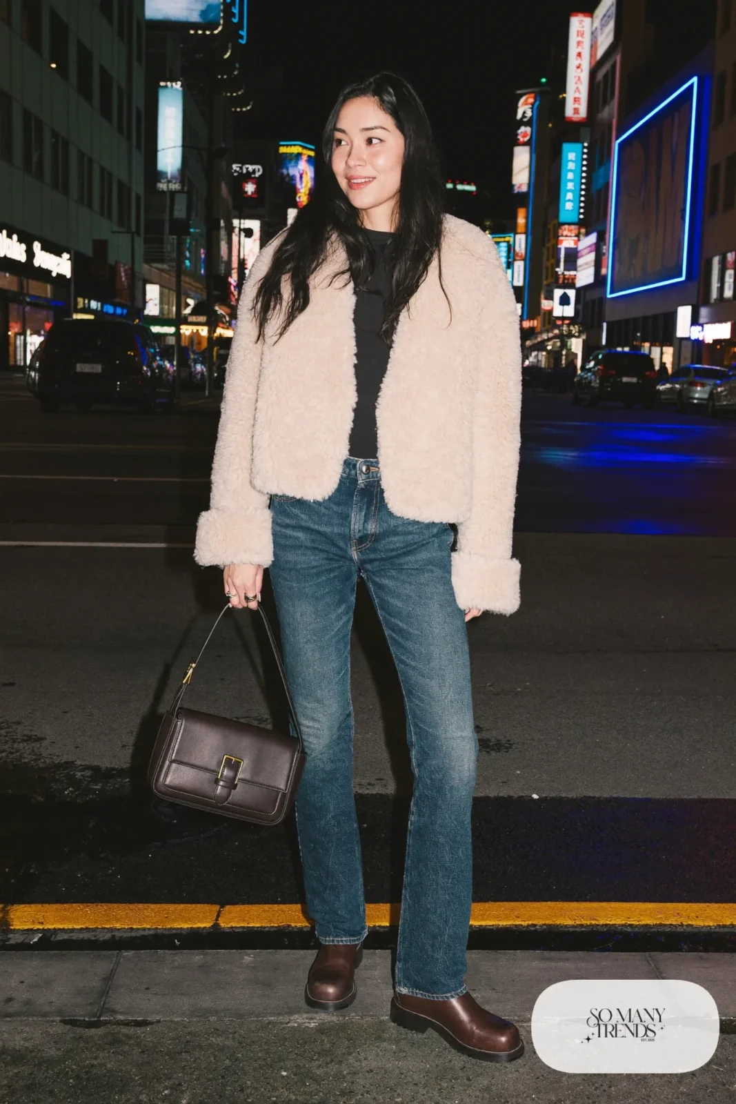 Woman in a cream cropped shearling jacket, black knit top, indigo jeans and brown boots in Las Vegas at night, outfit 22