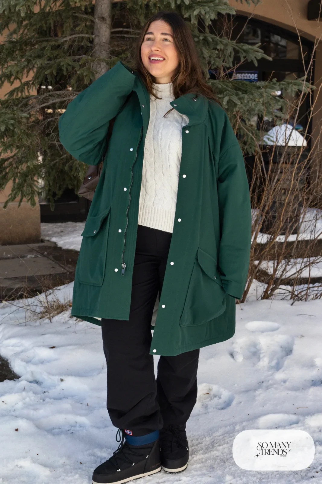 Look 10: forest green waterproof shell jacket over a cream knit sweater and dark pants with black snow boots, snowy woods