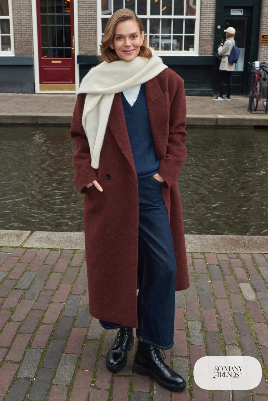 Woman in an oversized oxblood wool coat, navy knit sweater, wide-leg dark indigo jeans, black leather lace-up boots and ivory scarf on a canal street, outfit 1