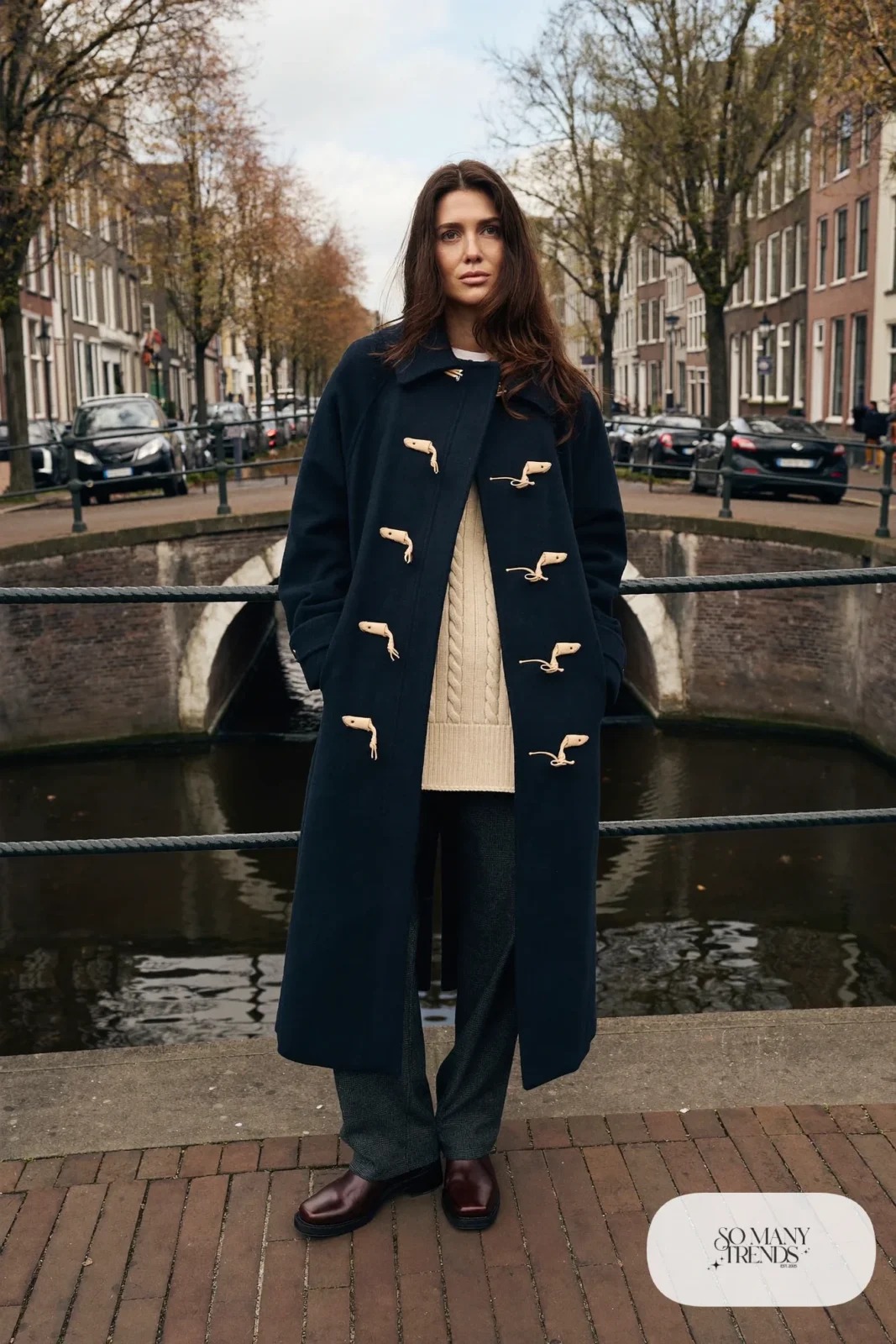 Woman in a navy toggle duffle coat, ivory cable knit sweater, straight gray wool pants and brown leather boots by a canal bridge, outfit 2