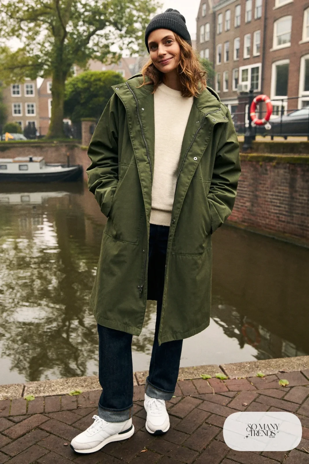 Woman in a forest green hooded parka, cream sweater, dark straight jeans, charcoal beanie and white leather sneakers on a canal quay, outfit 3