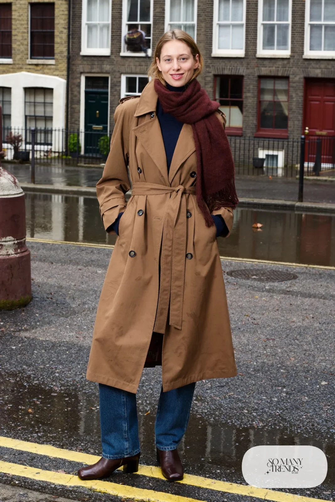 Woman in a belted camel trench coat, navy turtleneck, oxblood scarf, straight indigo jeans and brown leather boots on a canal street, outfit 4