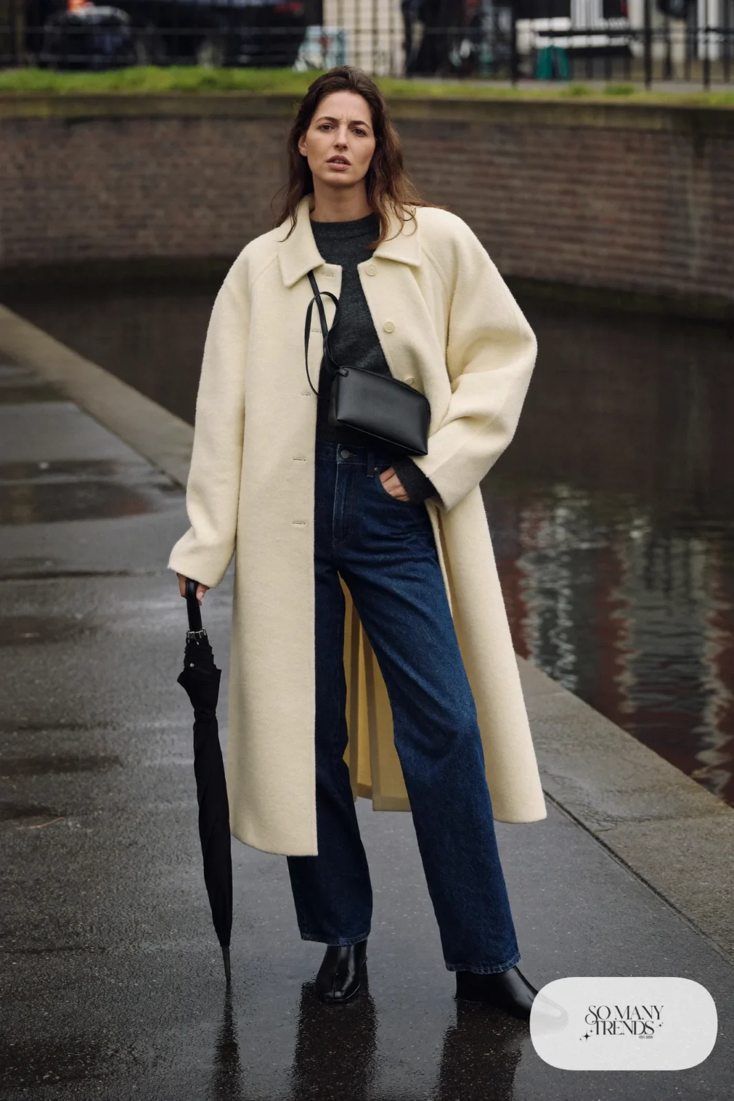 Woman in a long ivory wool coat, charcoal sweater, dark denim jeans, black leather boots and black crossbody bag on a canal street, outfit 6