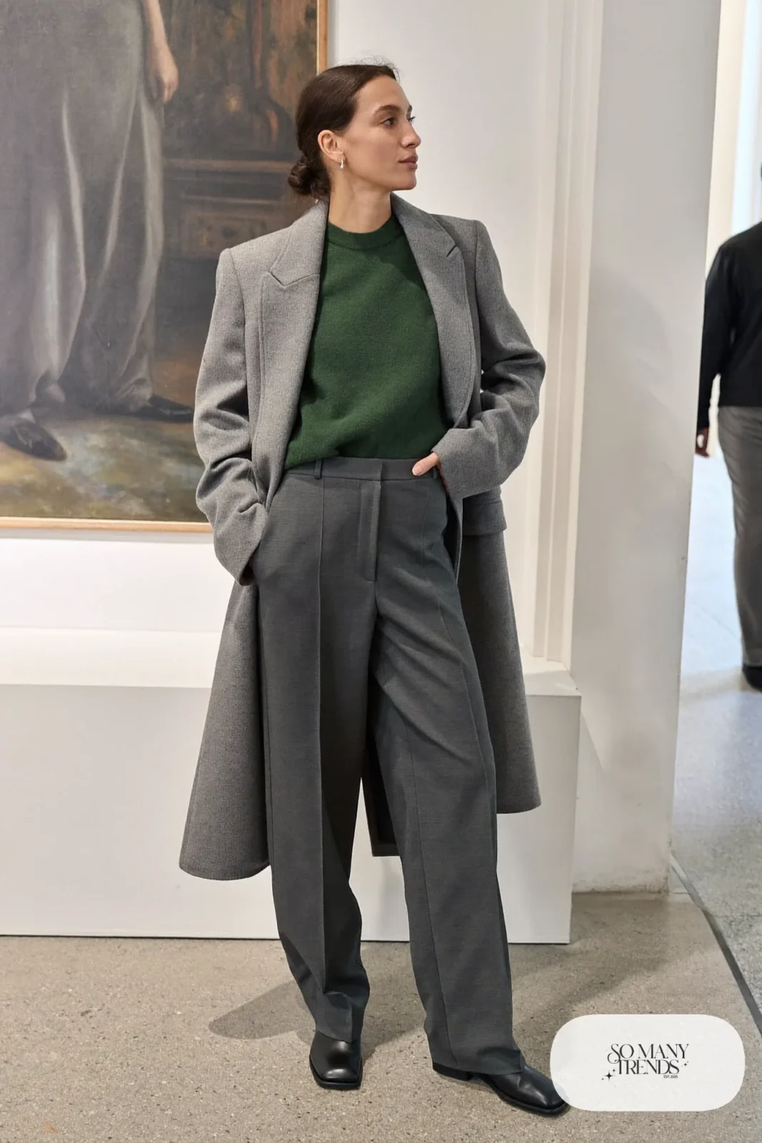Woman in charcoal tailored pants, forest green knit sweater, long gray blazer and black flat leather boots at a museum, outfit 11