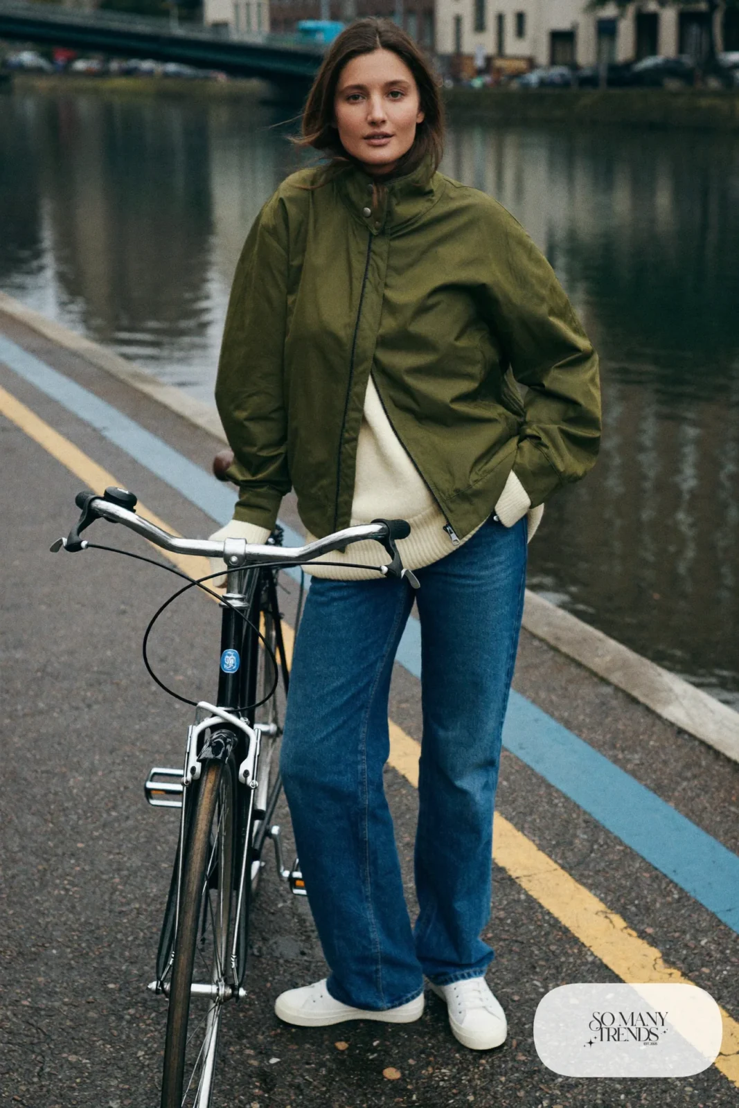 Woman in a boxy waxed cotton jacket, oxblood beanie, thick sweater, straight jeans and white sneakers by a bicycle on a canal street, outfit 14
