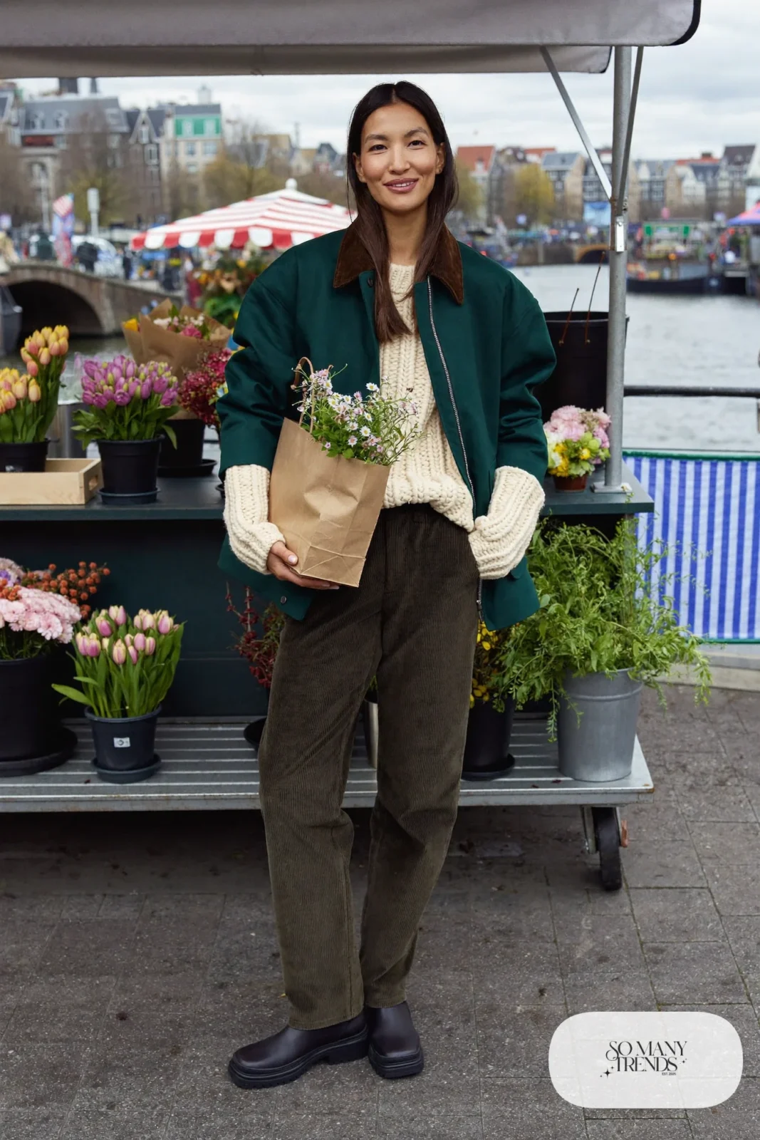 Woman in brown corduroy pants, cream wool sweater, green barn jacket, black rubber-sole boots and zip tote at a market, outfit 20