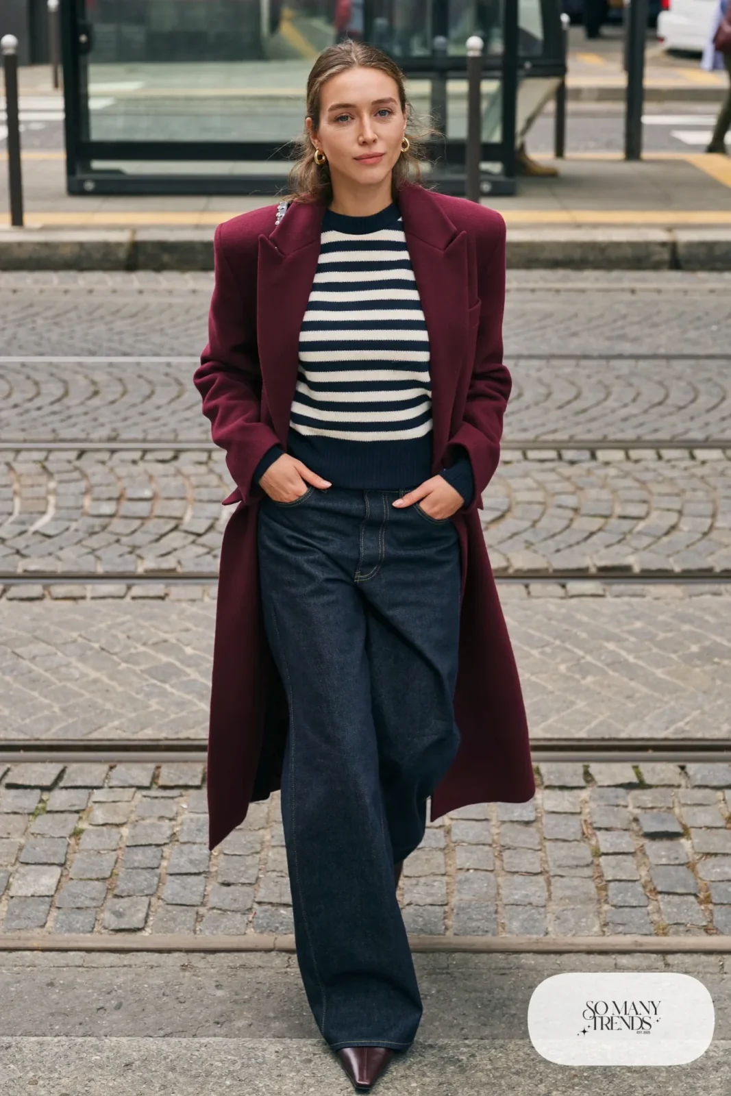 Woman in a navy and ivory striped sweater, oxblood coat, wide-leg dark jeans and ankle boots on a cobblestone street, outfit 24