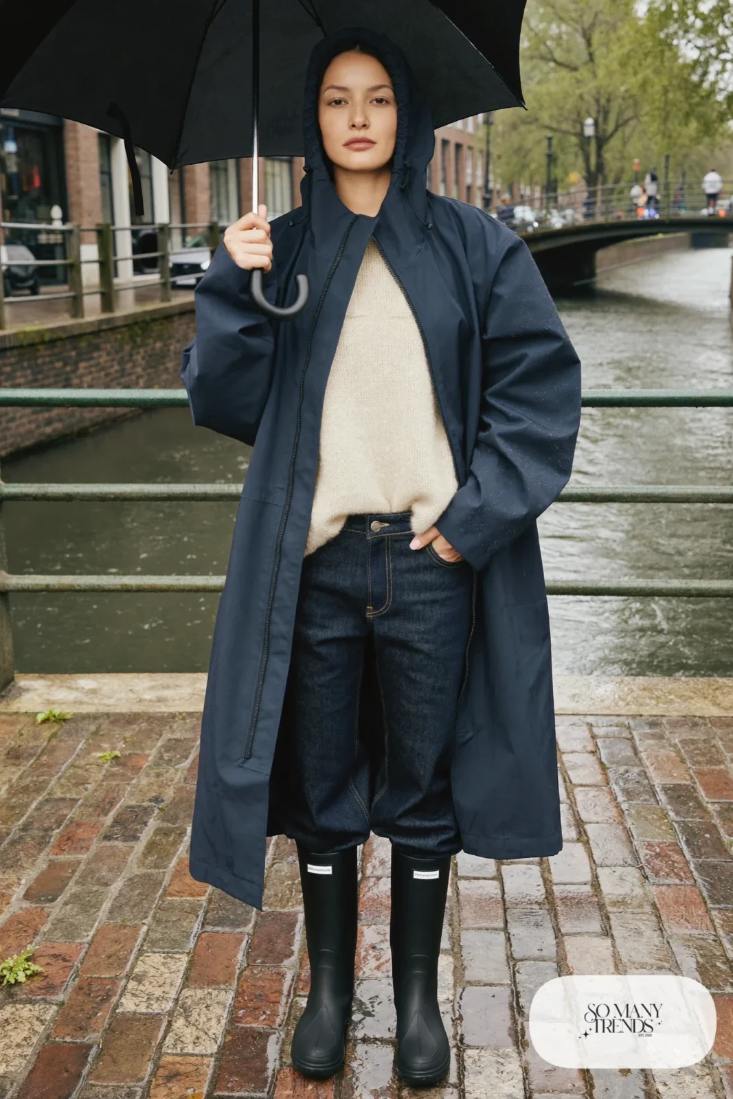 Woman in a navy long-line rain jacket, thick sweater, straight jeans and black waterproof boots in the rain on a canal bridge, outfit 25