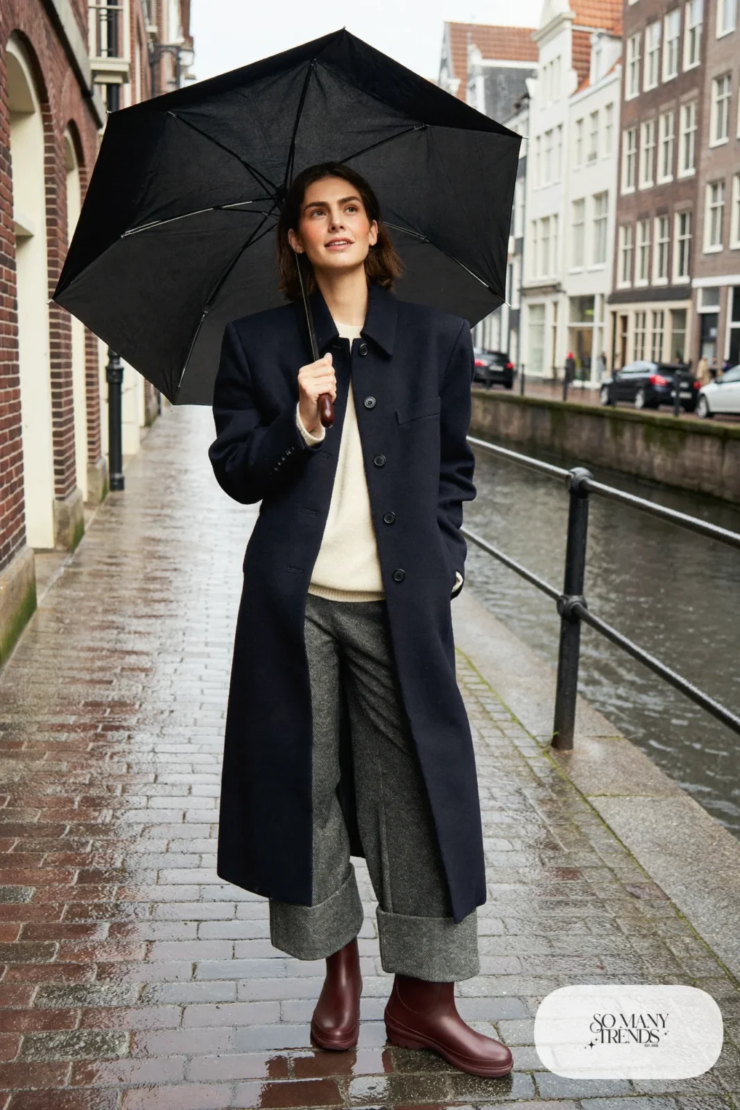 Woman in oxblood rain boots, gray wide-leg pants cuffed, cream sweater and navy coat walking on wet cobblestones, outfit 26
