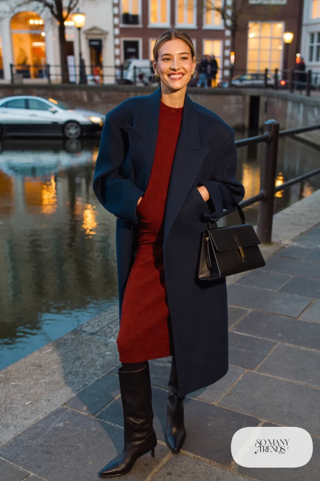 Woman in an oxblood knit midi dress, navy wool coat, black opaque tights, tall black boots and small black bag on a canal street at dusk, outfit 28