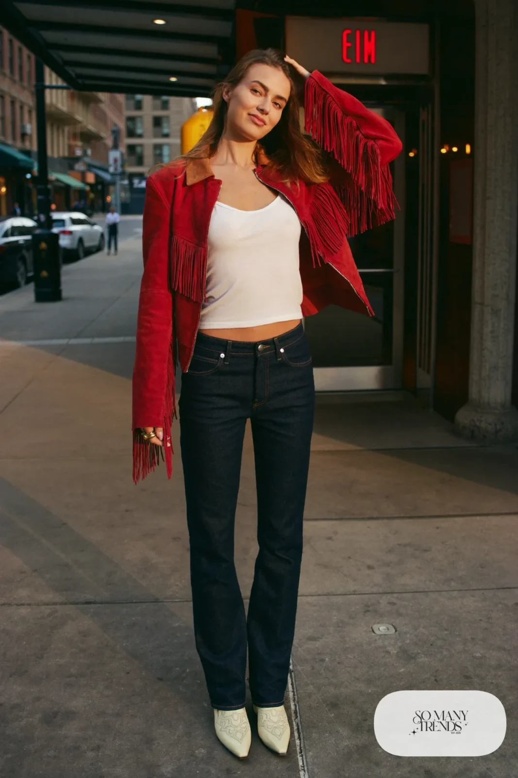 Woman in tomato red cropped suede fringe jacket, white tank, dark navy high-rise jeans and ivory pointed cowboy boots, outfit 1