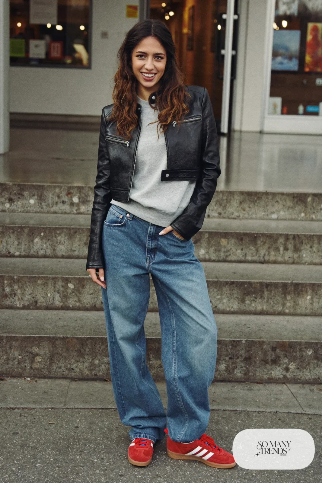 Woman in a black leather moto jacket, gray marl sweatshirt, baggy blue jeans and red Adidas Gazelle sneakers, outfit 6