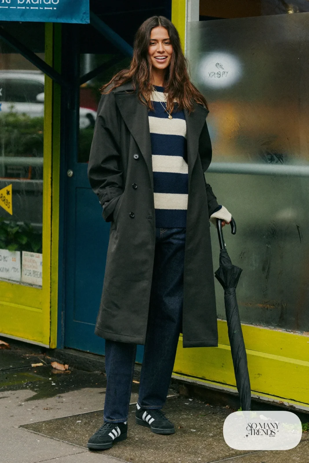 Woman in black trench coat, navy and cream striped sweater, dark straight jeans and black Adidas Campus sneakers, outfit 18