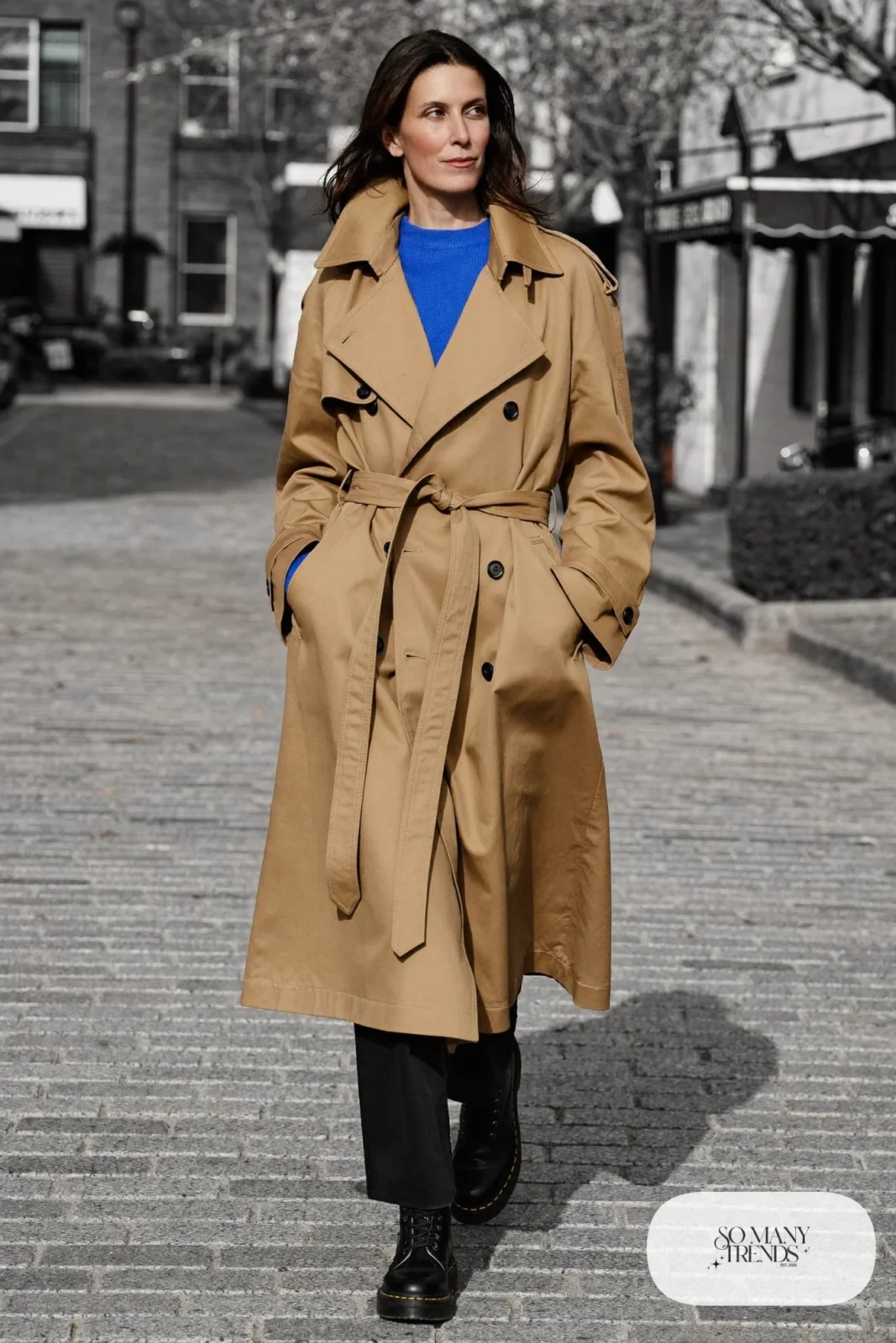 Woman in camel belted trench coat, cobalt knit sweater, black pants and black Doc Martens boots, outfit 16