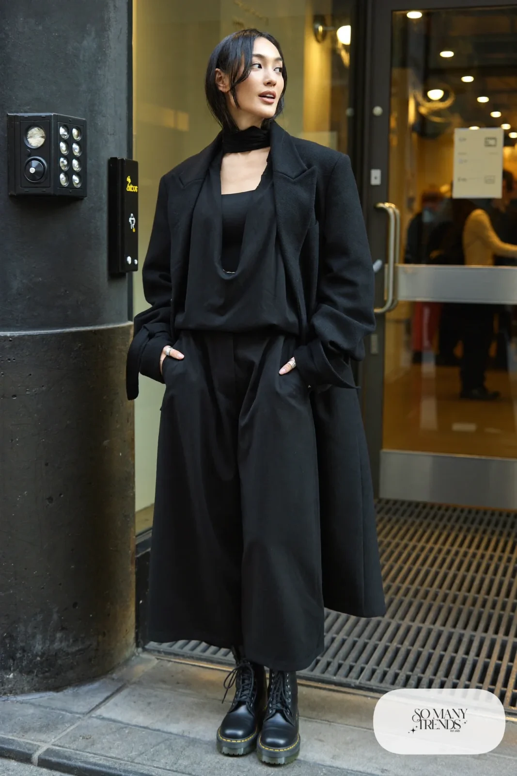 Woman in sheer black loose top over camisole, wide black pants, black Doc Martens boots and long black coat, outfit 23