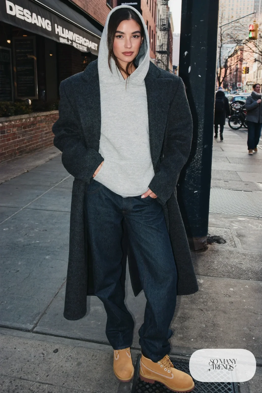 Woman in gray hoodie under charcoal tailored coat, dark jeans and wheat Timberland-style boots, outfit 18