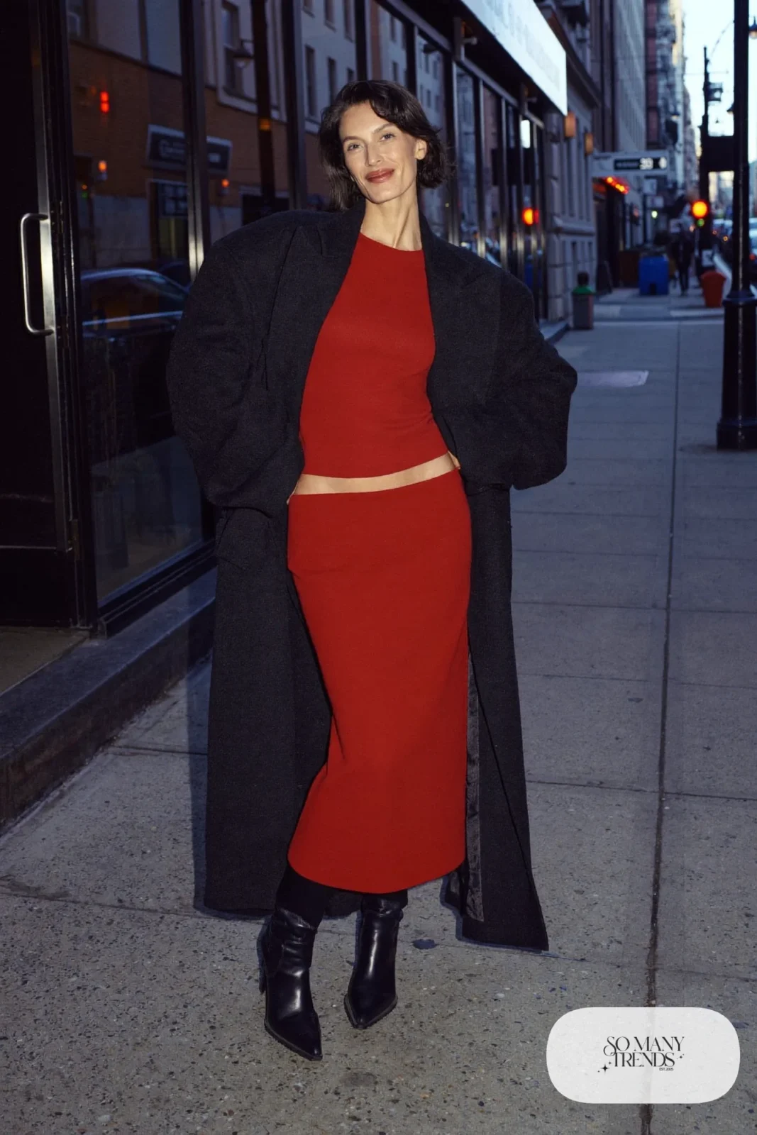 Woman in a red knit top and midi skirt set, black wool coat and black knee-high boots, look 22