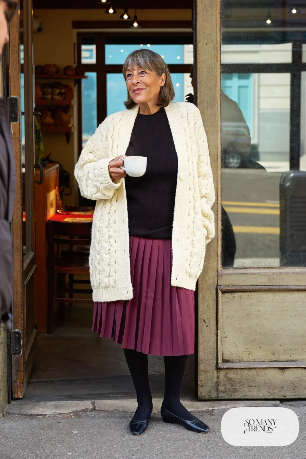 Woman over 50 in an ivory cable-knit cardigan, plum pleated midi skirt, black opaque tights and black ankle-strap flats, Christmas outfit 3