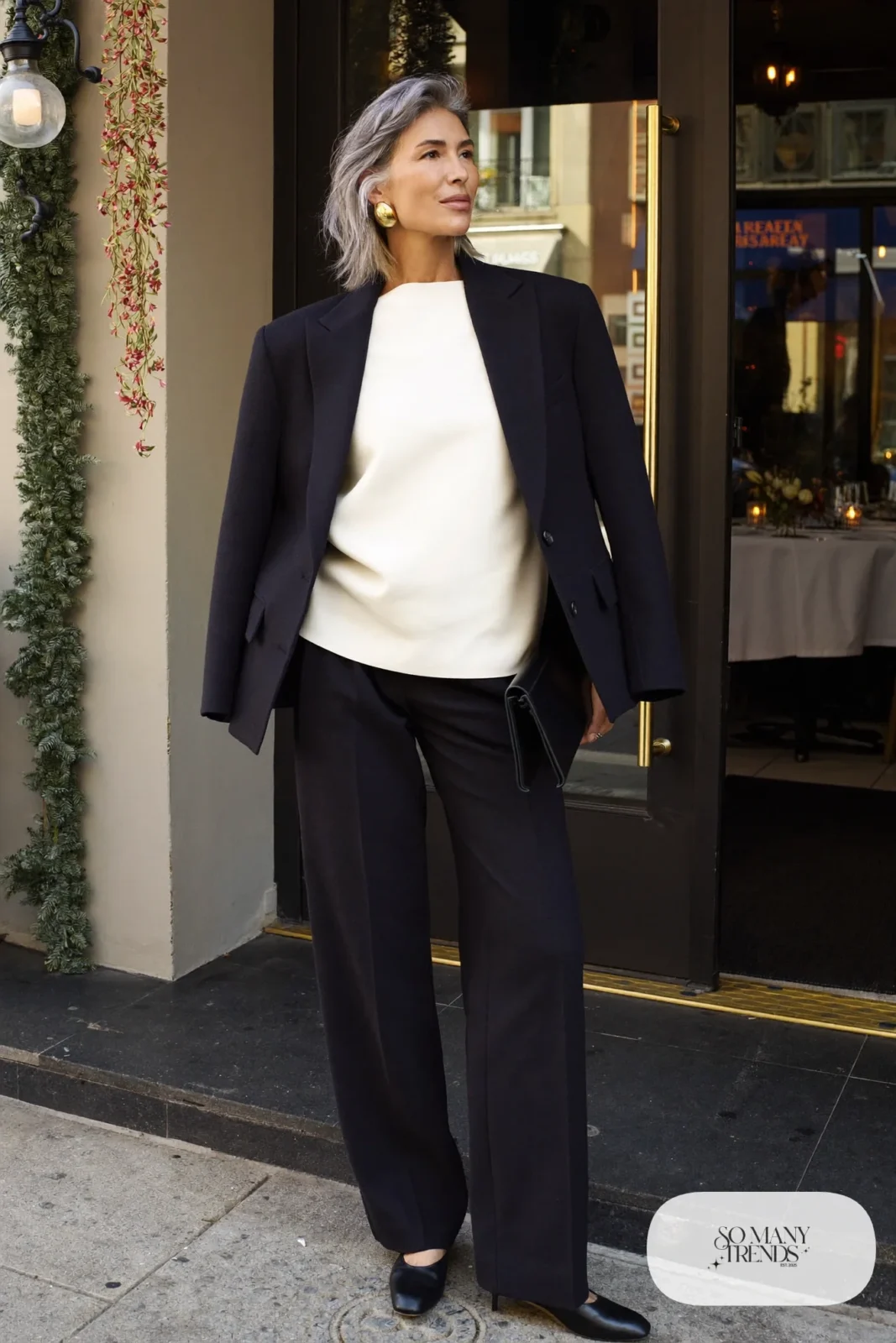 Woman over 50 in a navy tailored blazer, matching wide-leg pants, ivory shell top, gold hoops and black pumps, Christmas outfit 13