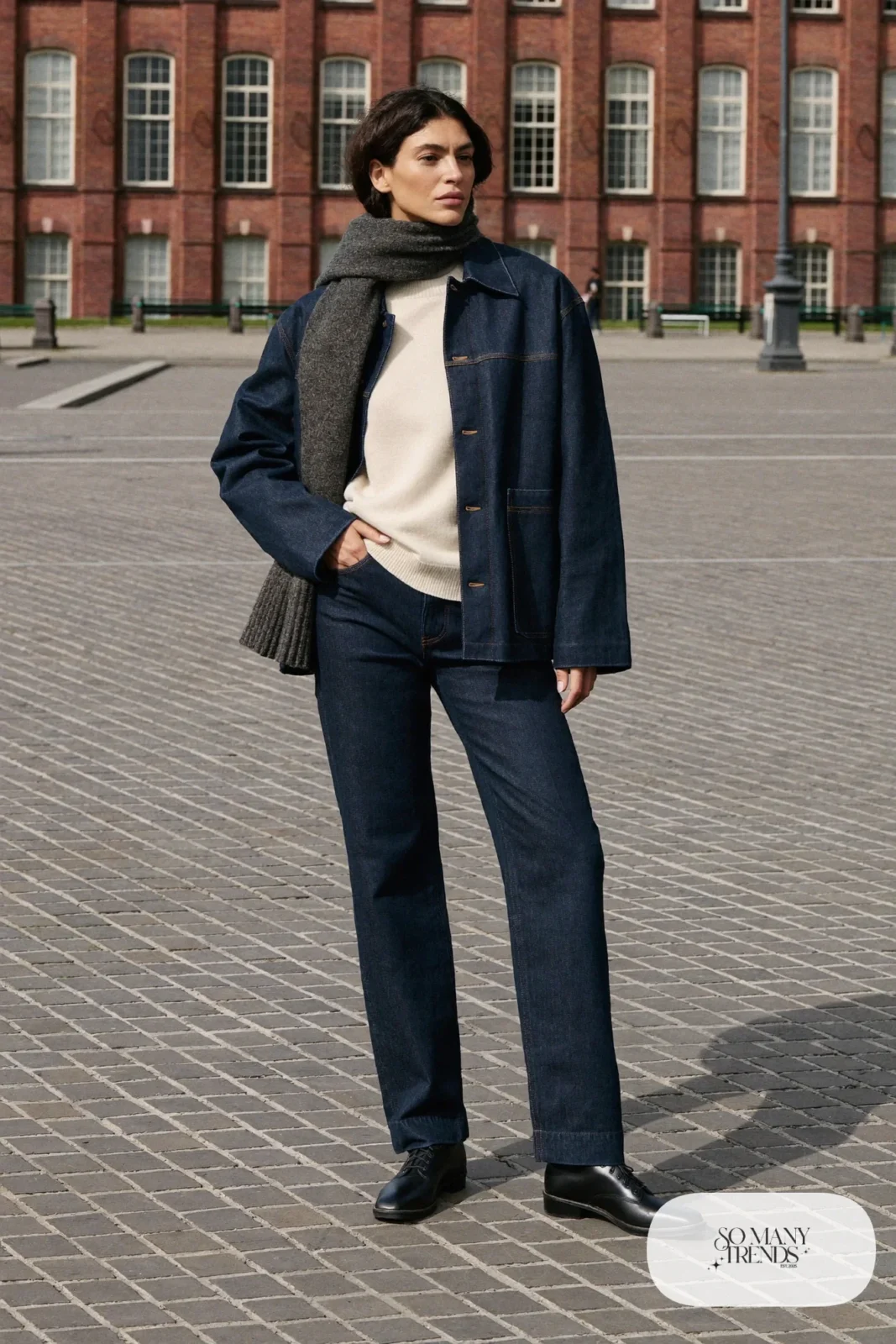 Woman in a boxy dark denim jacket, ivory knit sweater, dark straight denim jeans, black boots and gray scarf on a museum square, outfit 9