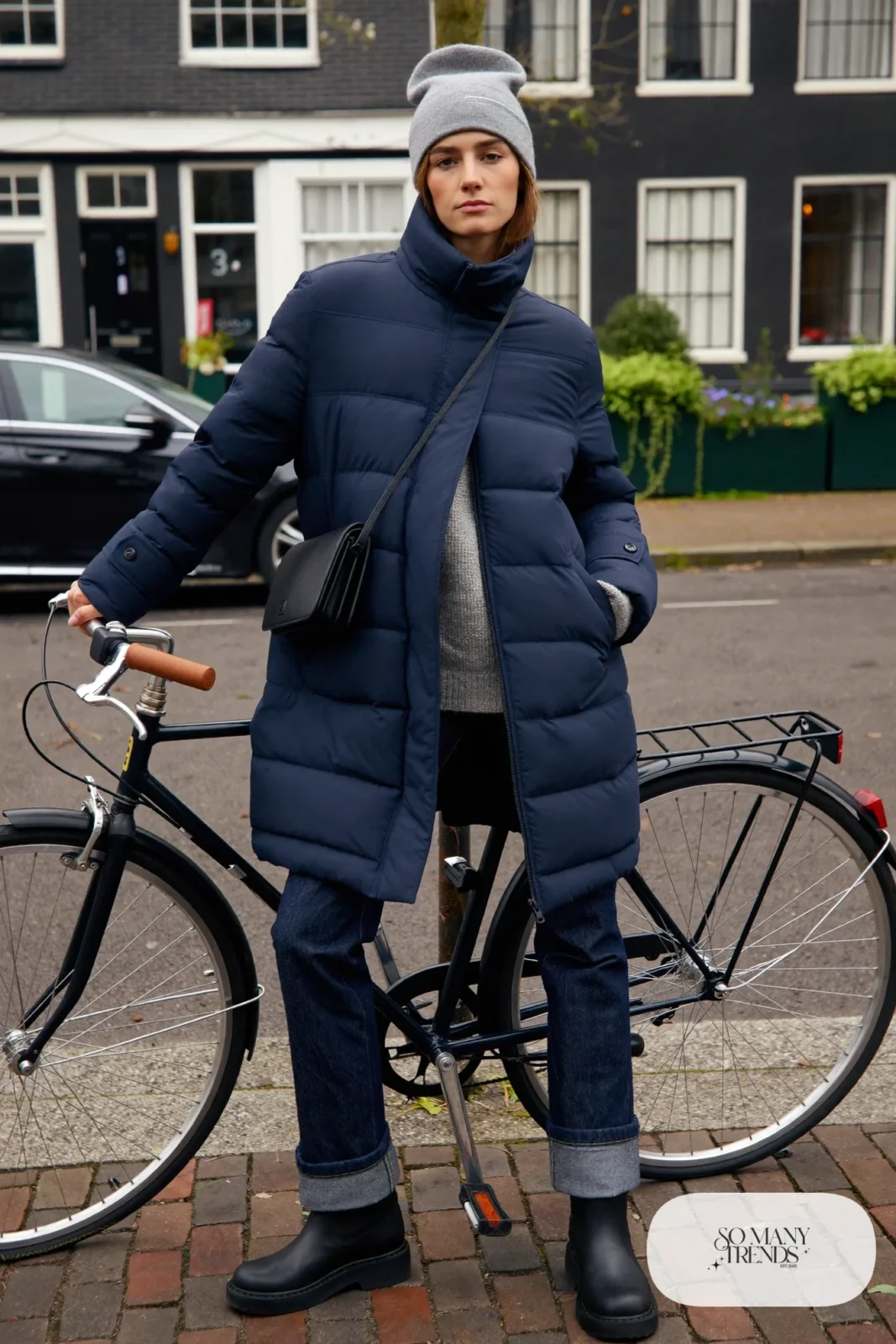 Woman in a navy mid-length padded coat, dark straight jeans, waterproof ankle boots, gray beanie and crossbody bag beside a bicycle, outfit 13