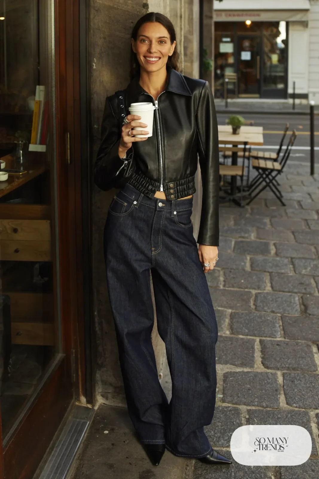 Woman in a cropped black leather jacket, long oxblood sweater, high-rise wide dark jeans and pointed black boots outside a cafe, outfit 21