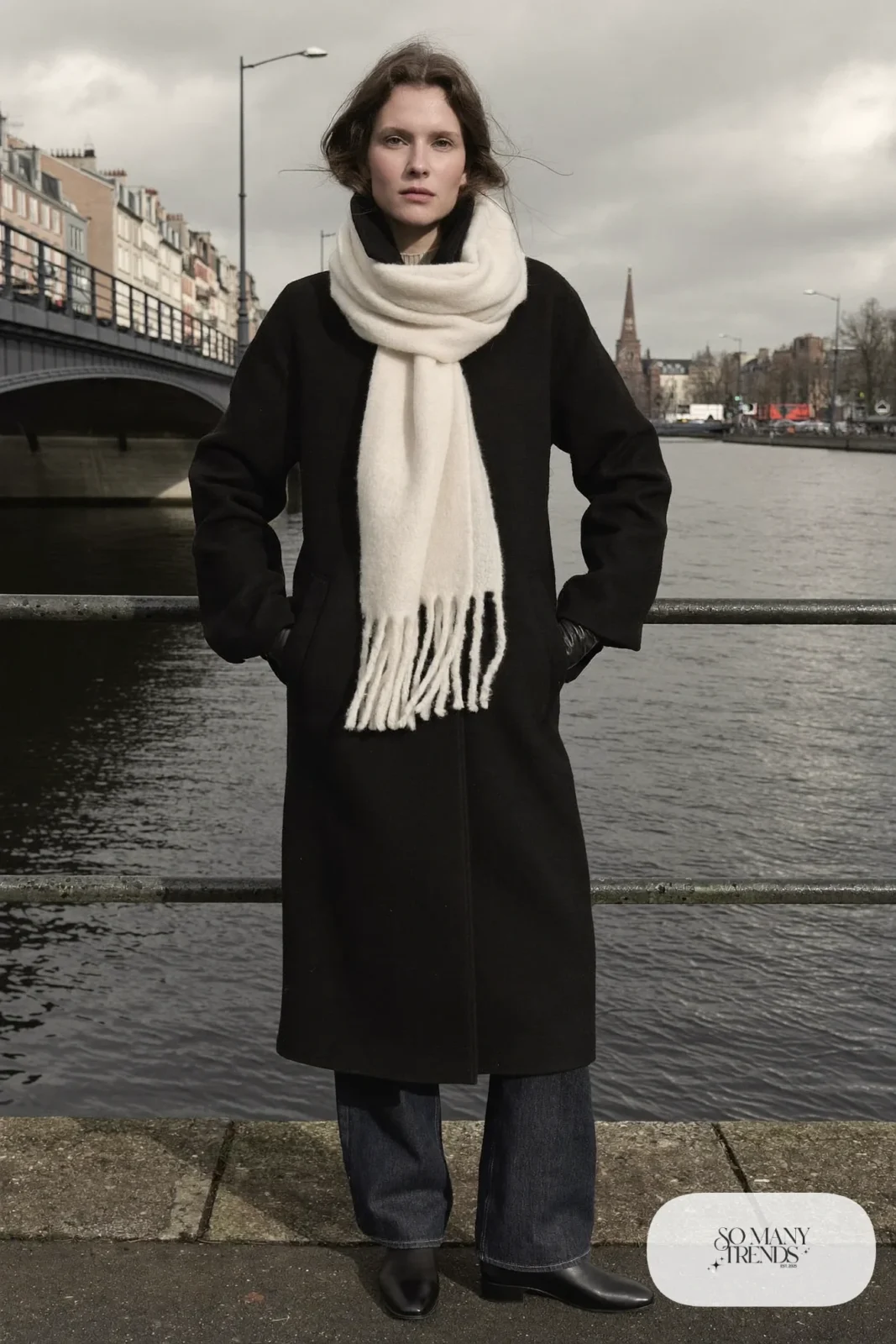 Woman in a long black wool coat, ivory scarf wrapped twice, dark jeans, black leather gloves and flat black boots by a canal, outfit 27